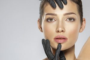 A imagem mostra o rosto de uma mulher branca com cabelos castanhos. Ela olha para frente e possui luvas pretas em suas mãos – uma delas está tocando a sua testa e a outra está posicionada sobre seu queixo.