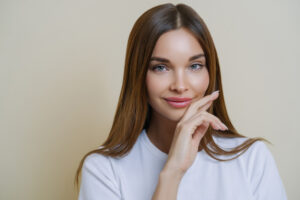 A imagem mostra uma mulher branca, com olhos azuis, cabelo castanho e liso. Ela veste uma camiseta branca com gola redonda. Com uma das mãos ela apoia a área lateral do queixo. A modelo está sorrindo e o fundo da imagem é amarelo claro.