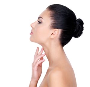 A imagem mostra uma mulher branca, com cabelos pretos presos em um coque. Ela está virada de perfil, com o rosto levemente elevado. Com uma das mãos ela toca o pescoço. O fundo da fotografia é branco.