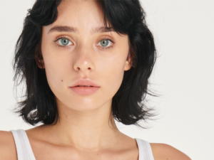 A imagem mostra uma mulher jovem, com pele clara e cabelos pretos na altura do pescoço. Ela tem olhos azuis e olha fixamente para frente. A modelo veste uma regata com alças brancas. O fundo da imagem é cinza claro.